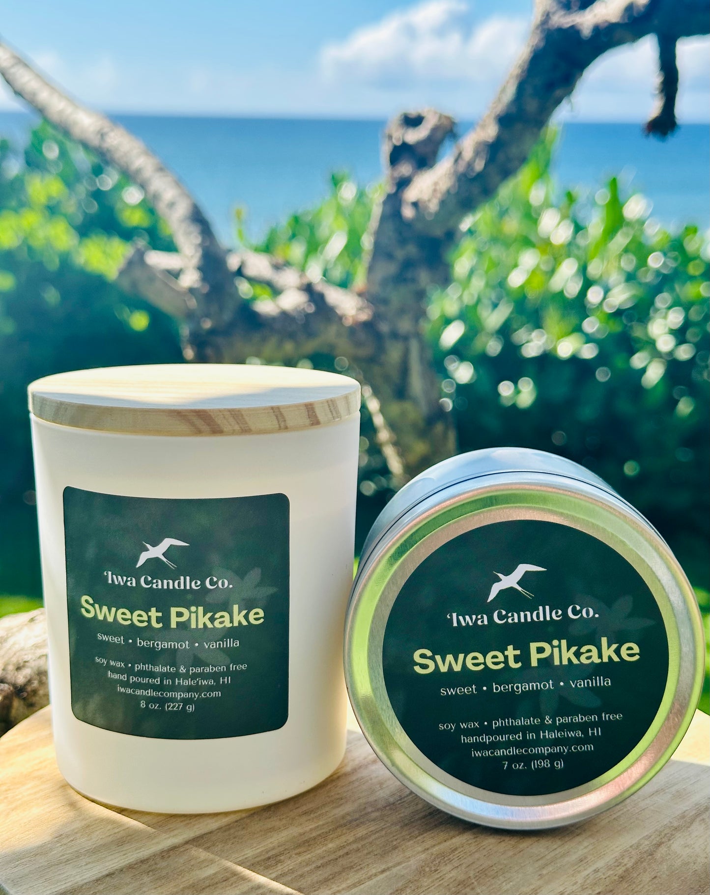 Two 'Iwa Candle Co.' Sweet Pikake scented candles, one in a white jar with a wood-grain lid and one in a round tin, displayed on a wooden surface with a blurred outdoor ocean and tree background.