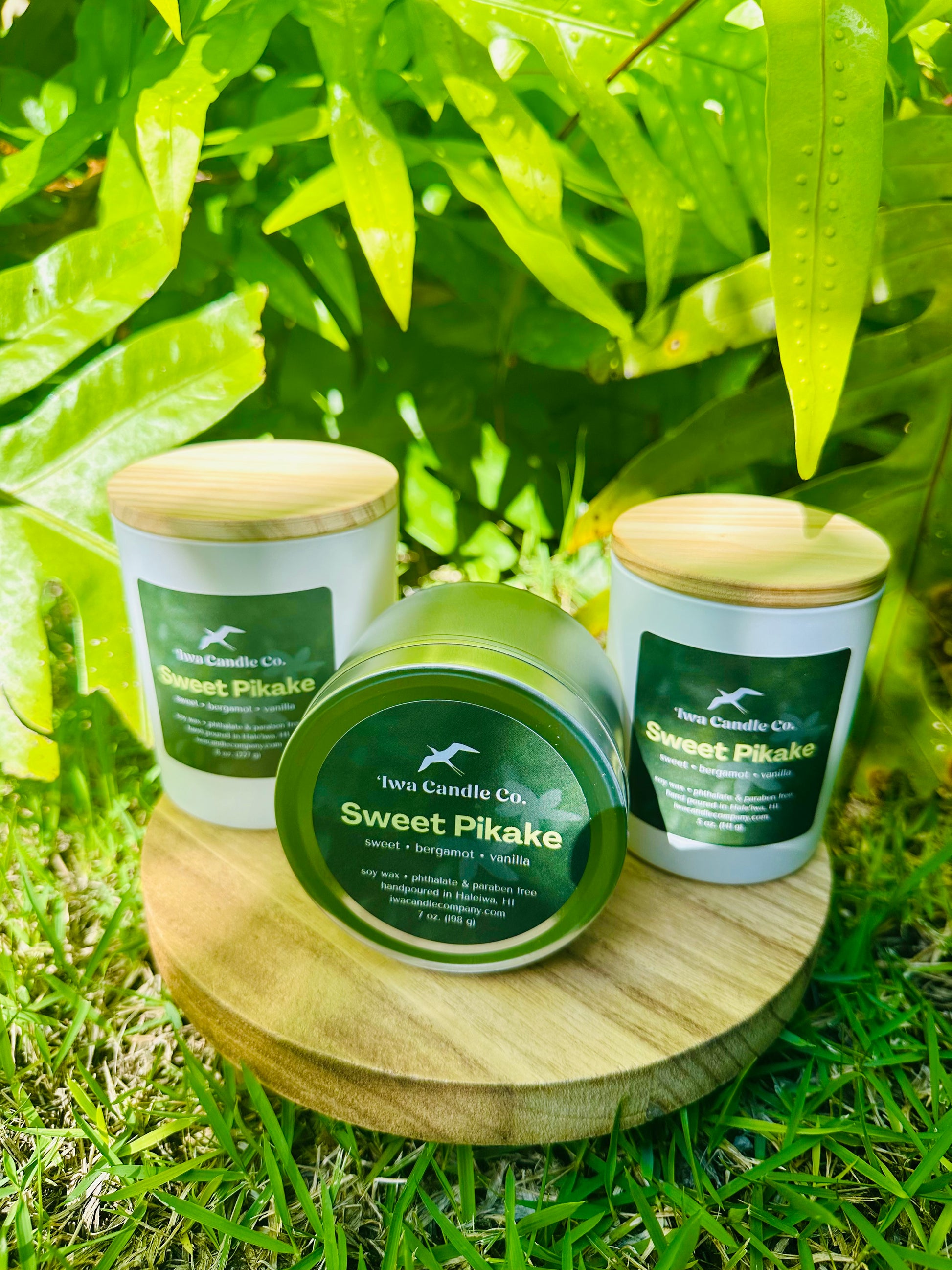 Three scented candles are displayed outdoors on a round wooden tray, surrounded by lush green leaves and grass.